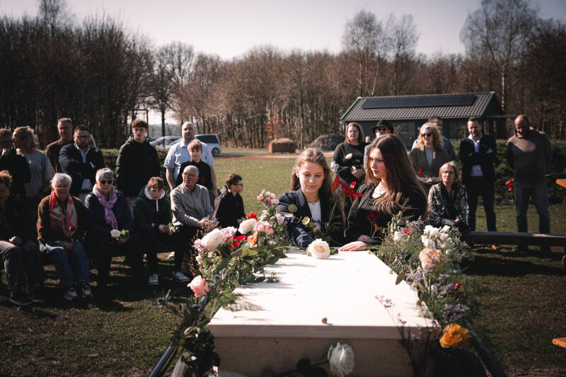 Nabestaanden staan samen rondom een lichte, met veldbloemen versierde kist in de natuur.