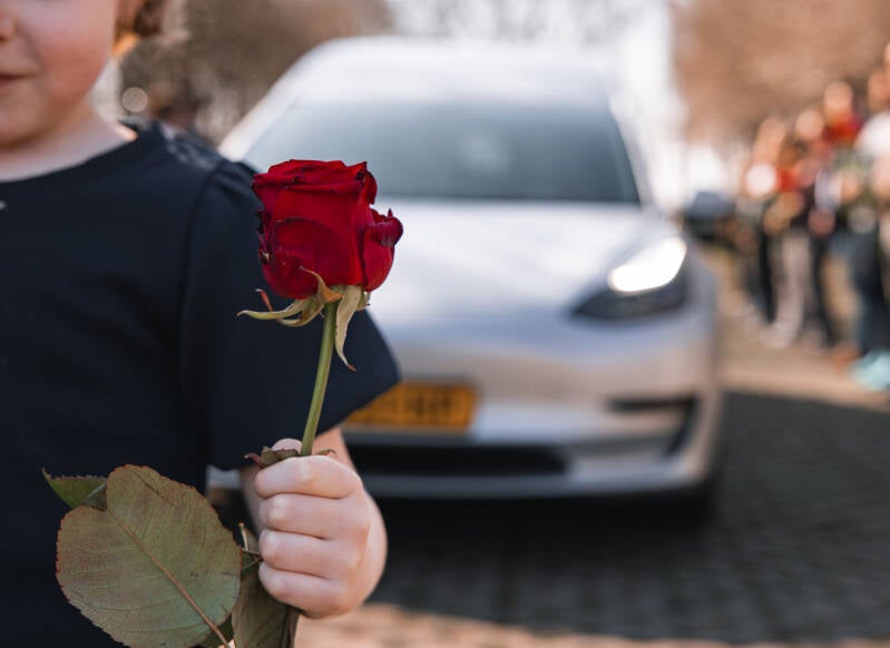 Detailfoto van een hand met een rode roos, met een moderne Tesla rouwauto op de achtergrond.