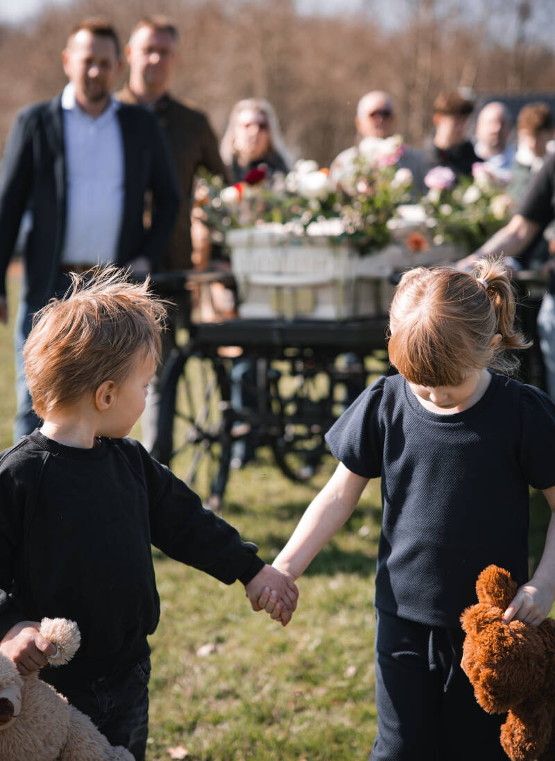 Twee jonge kinderen die hand in hand lopen bij een uitvaartkist in de natuur.