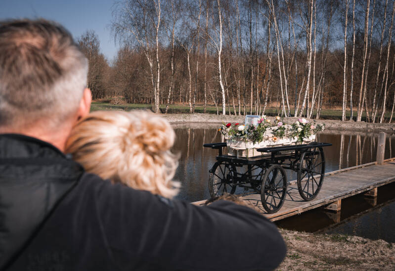 Nabestaanden die elkaar omhelzen en troosten bij het water, met de rouwkoets op de achtergrond.