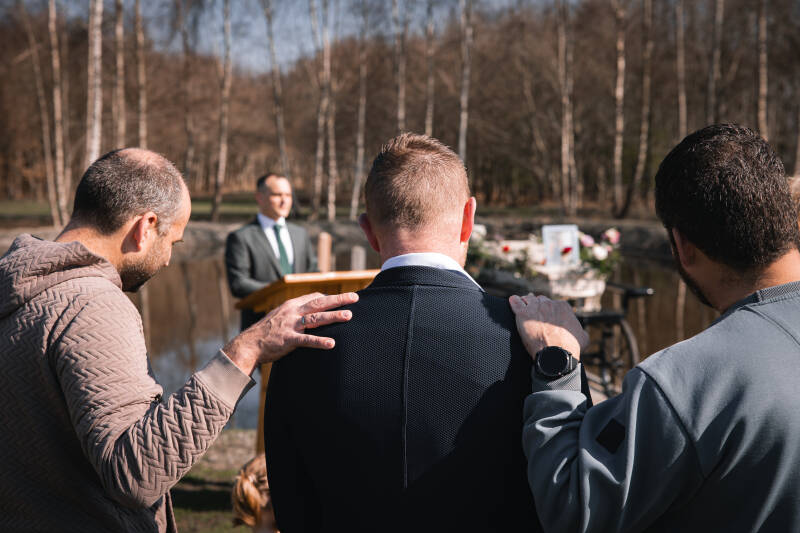 Uitvaartfotografie van drie mannen die elkaar troostend vasthouden bij een natuurbegrafenis.