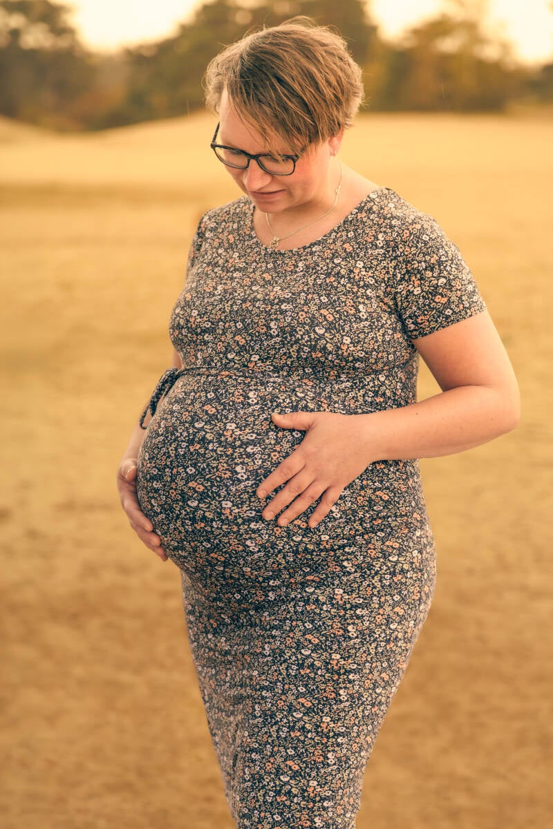 Intieme zwangerschapsfotografie op de zandverstuiving tijdens het gouden uur. Een zwangere vrouw houdt liefdevol haar buik vast, omhuld door warm avondlicht. High-end lifestyle fotografie door Mark Nederhoed.