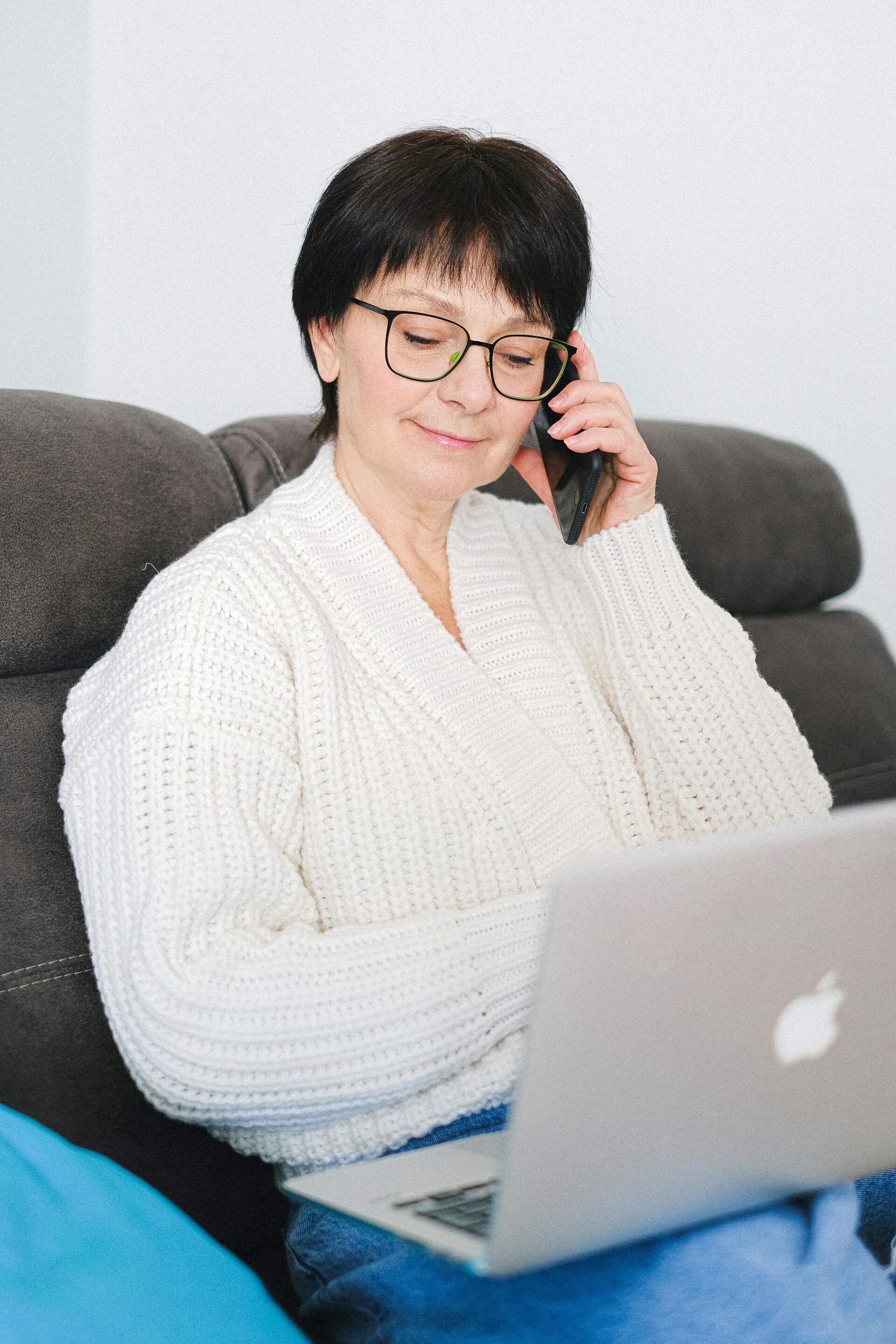 Smiling older woman in a white knit sweater talking on a cell phone while using her laptop
