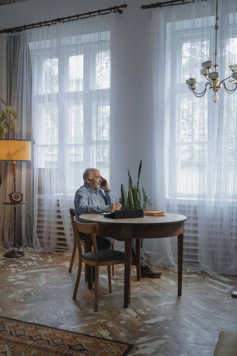 Older man sitting at a table in a bright living room talking on the phone