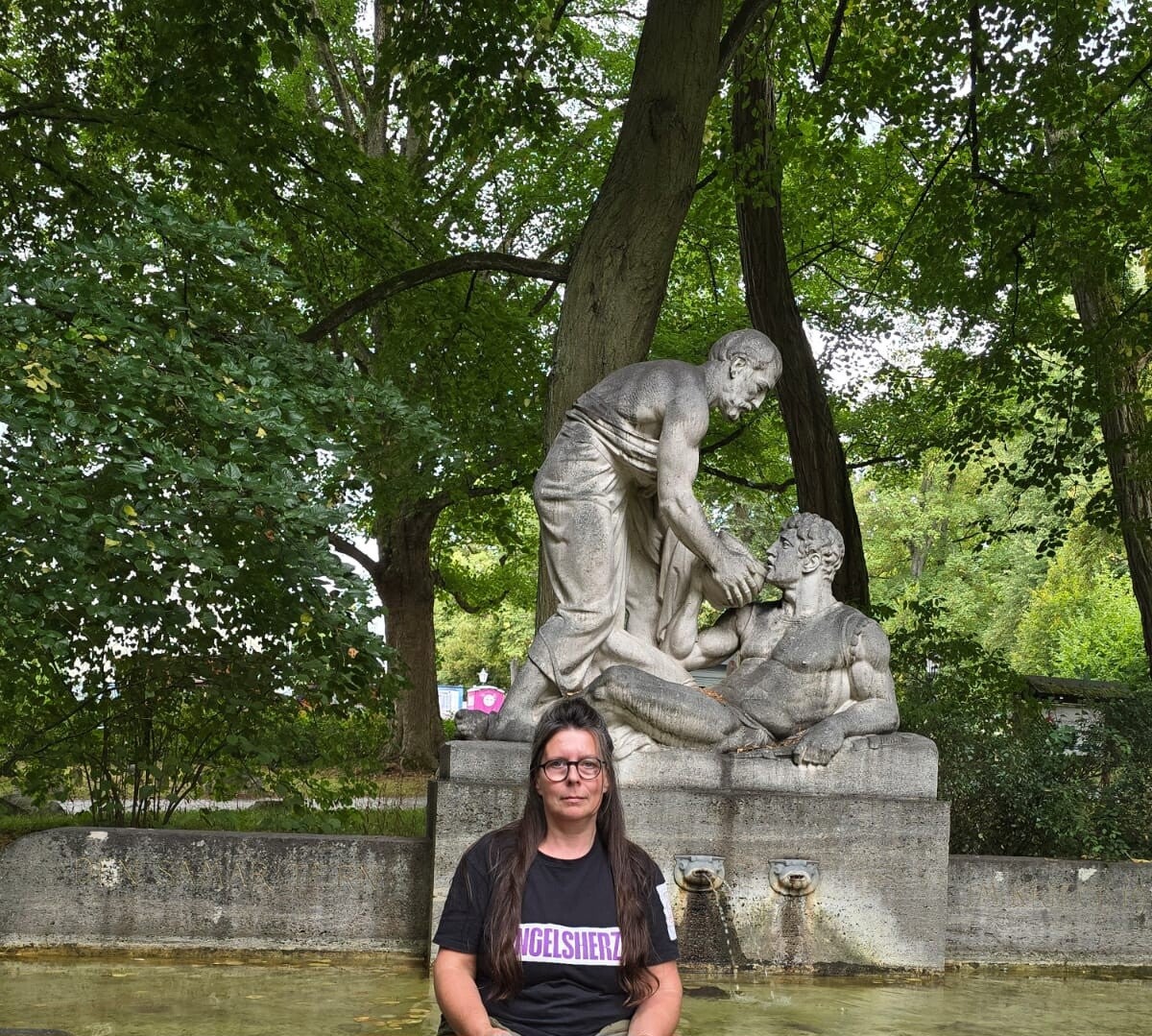 Tanja von Engelsherz sitzt vor dem steinernen Samariterdenkmal im grünen Kurpark von Bad Homburg.