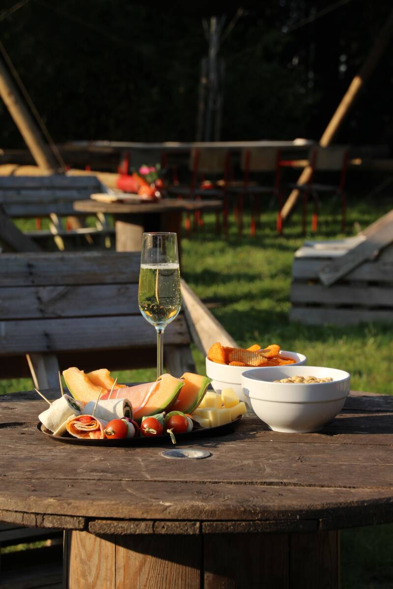 Een gazon met houten tafel gevuld met een glaasje bubbels en hapjes en in de verte een bbq