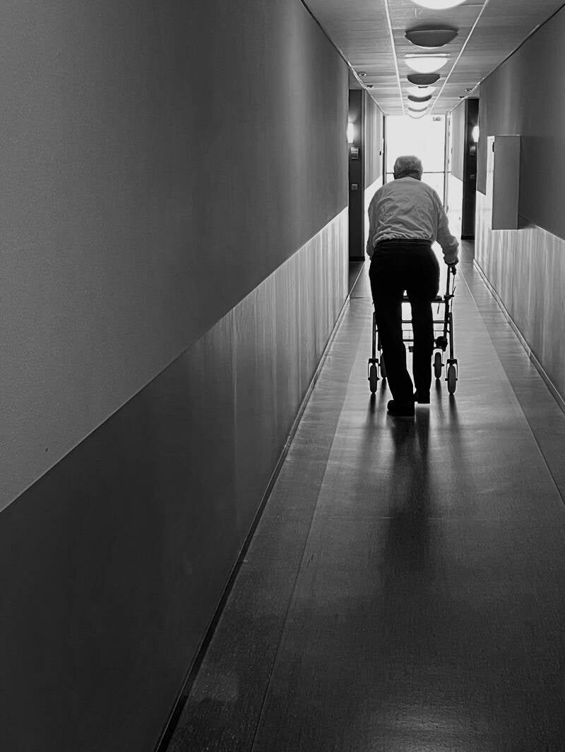 Een zwart wit foto in de gang van een woonzorgcentrum en in die gang loopt een man met een rollator