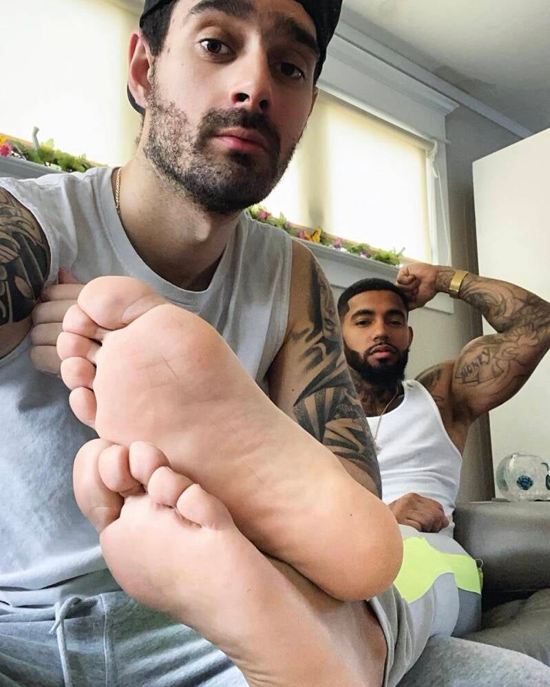 Guy holds another man's bare feet in his lap showing off fat toes, smooth soles.