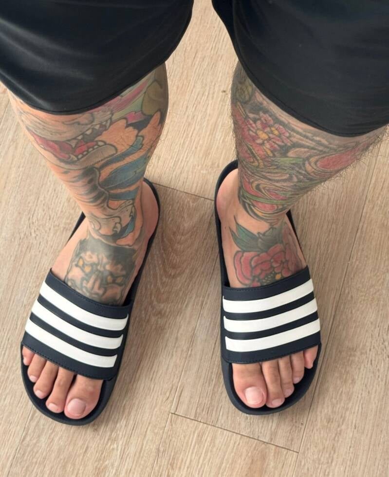Tattooed legs of a man wearing slides as he shows off top of his toes.