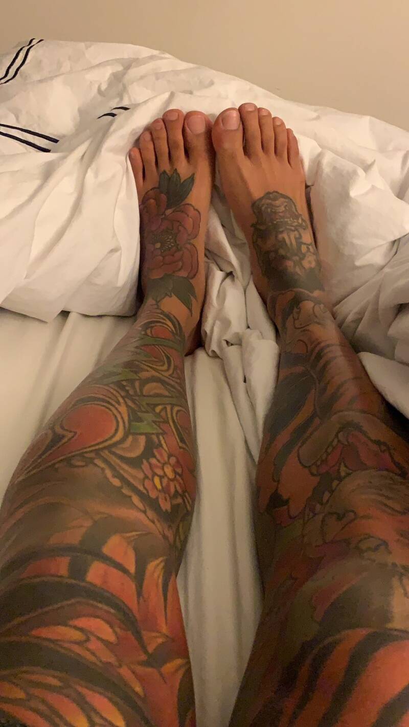 Big feet man showing off groomed masculine feet and tattooed legs while lying in bed.