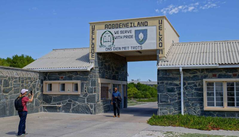 A white woman is taking a picture of a black male in front of the entrance of Robben Island Prison, South Africa