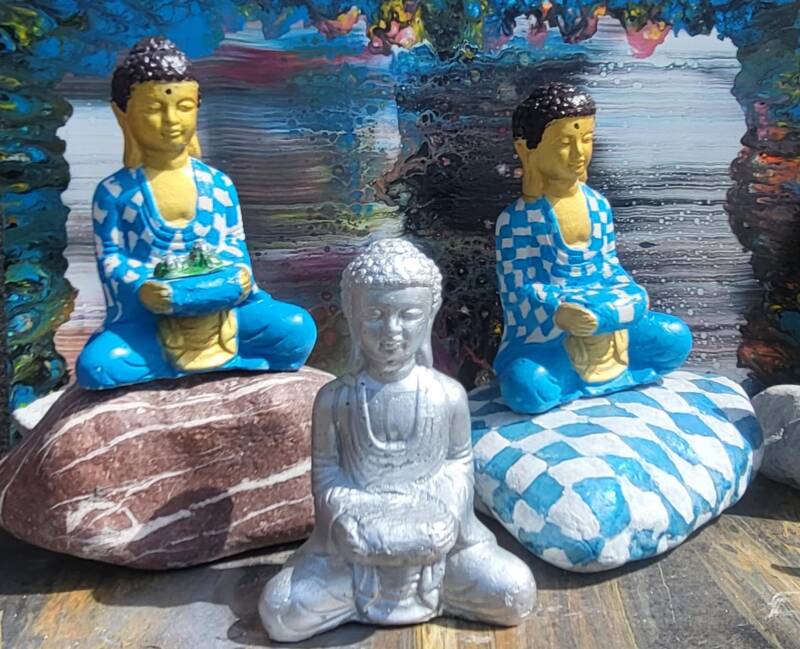 Bayerische Buddhas in weiß und Blau auf Steinen sitzend mit Spray Paint Bild im Hintergrund in blau rot weiß grün Lila bunte Streifen