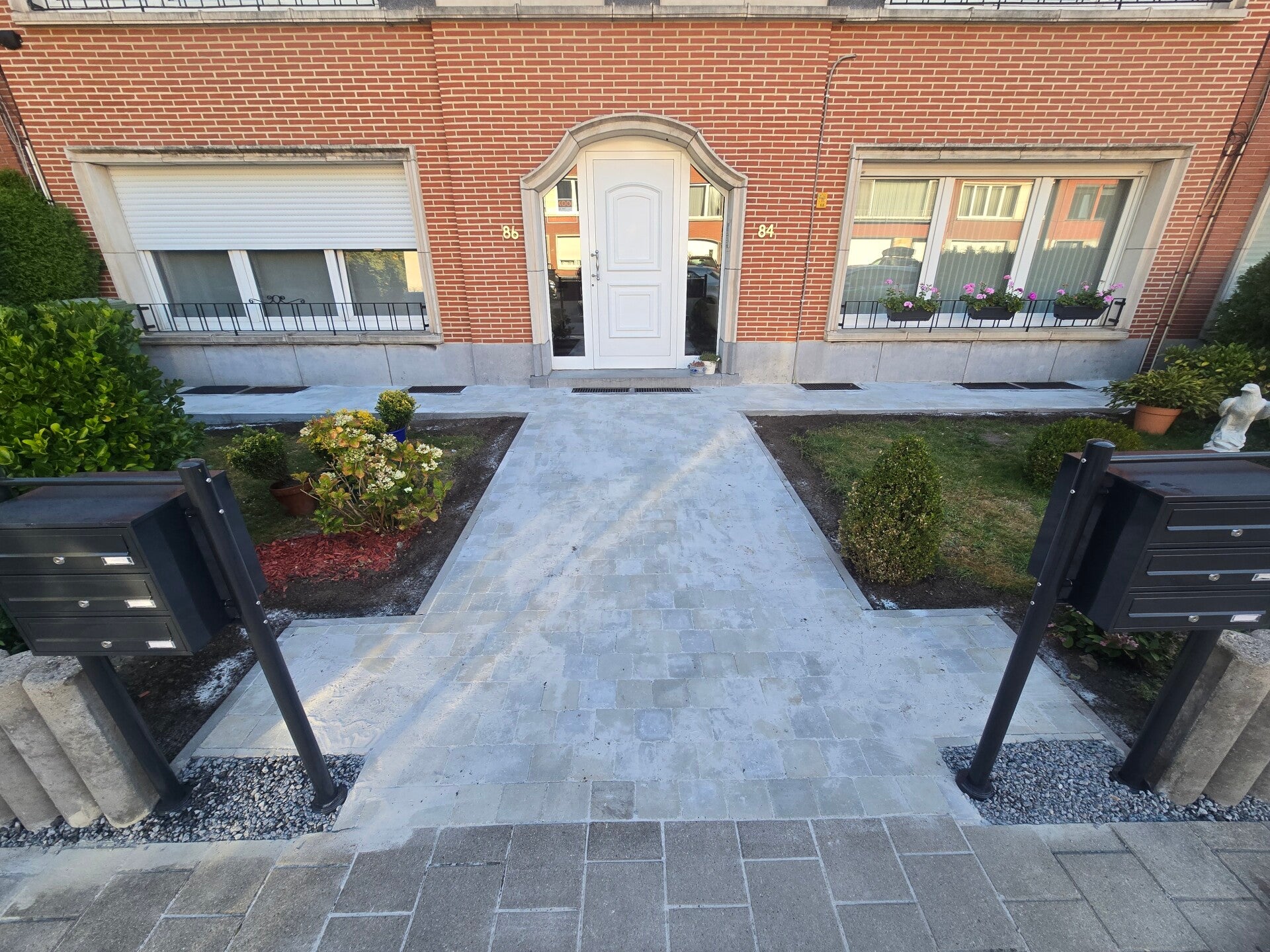 Nieuw aangelegd toegangspad in grijze klinkers naar de voordeur van een woning, symmetrisch uitgezet met donkere boordstenen.