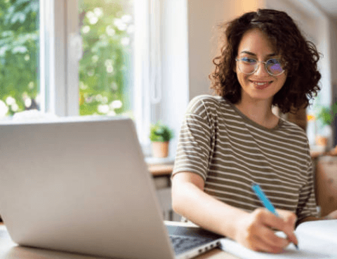 Confident student smiling at a laptop and taking notes during successful advanced A-Level French grammar tuition classes.