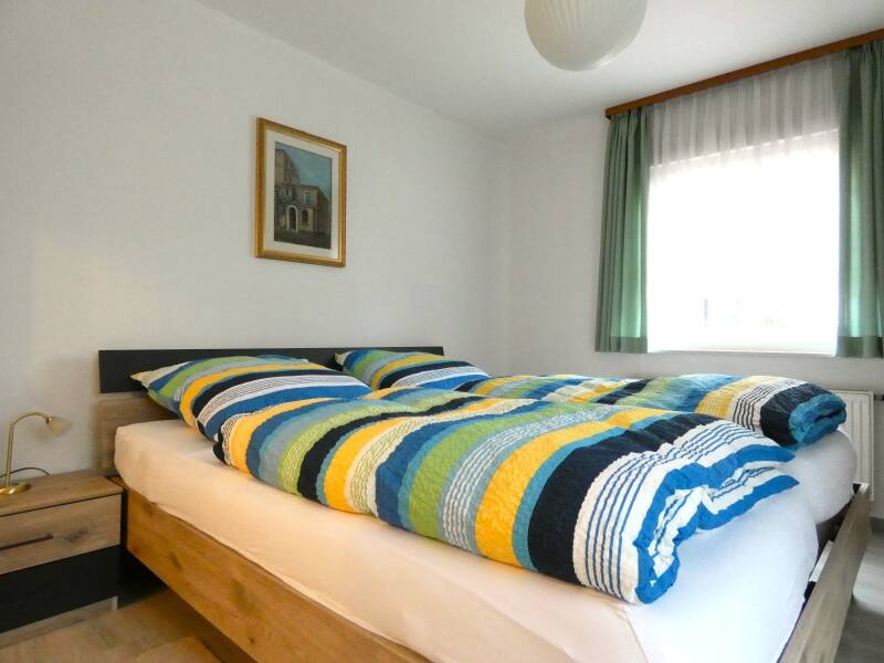Das lichtdurchflutete Doppelzimmer in der Ferienwohnung Klingel mit fröhlich gestreifter Bettwäsche einem mediteranen Bild und Fenster mit grünem Vorhang