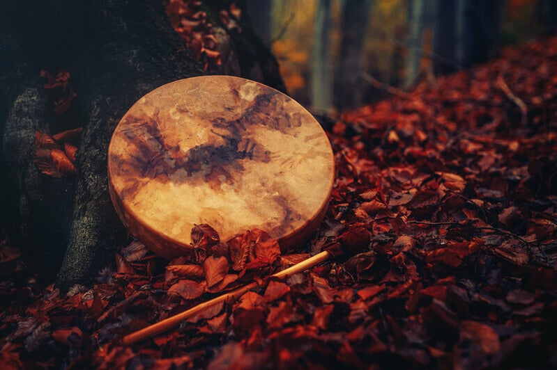 Runde Rahmentrommel mit Fellbespannung lehnt an Baumstamm, liegt auf Boden mit roten Herbstblättern, daneben ein Schlägel, Kontext Blog Mediumship & Research Institut für Medialität & Afterlife Forschung.
