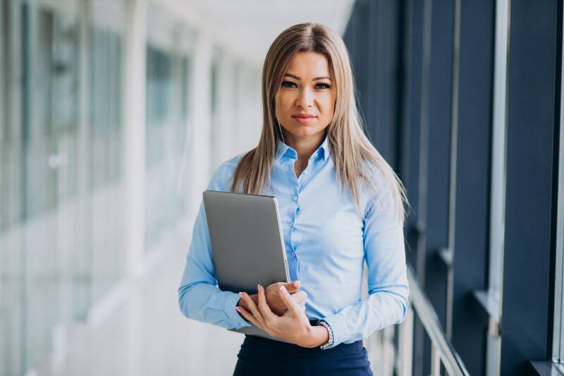 Junge Geschäftsfrau steht in einem Büroflur und hält einen Laptop vor der Brust, im Kontext mit Jenseitskontakte Jana Lorenz.