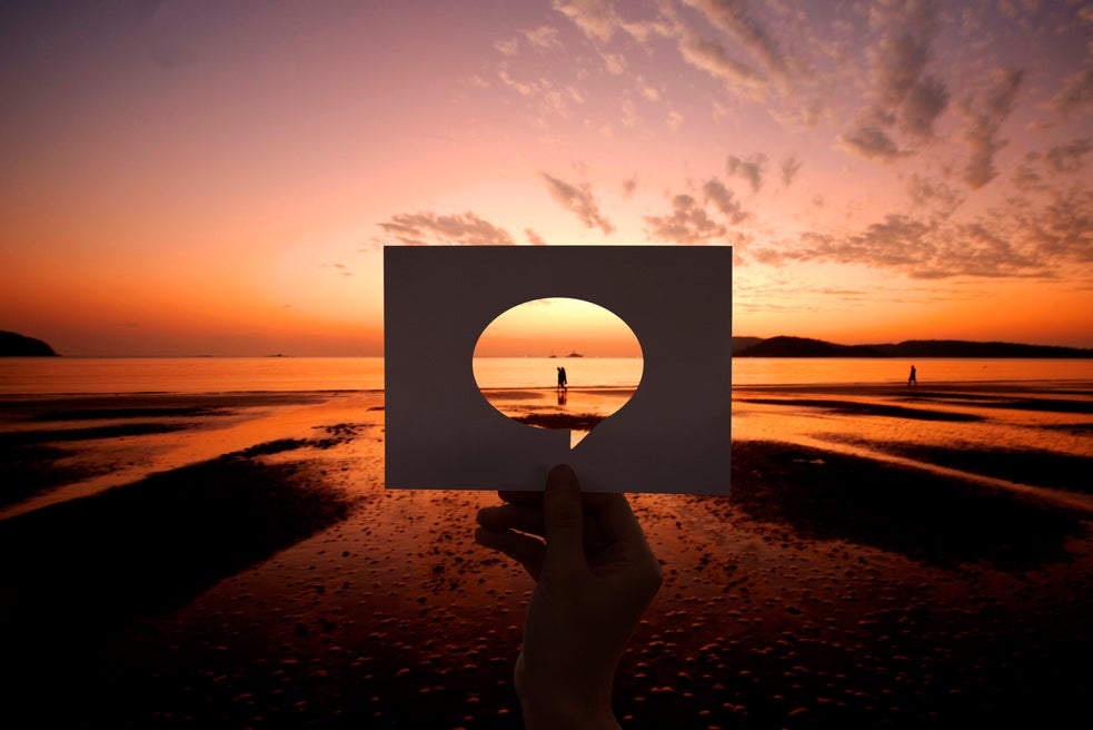 Hand hält rechteckiges Papier mit ovalem Loch vor Sonnenuntergang am Strand, Person im Hintergrund sichtbar, Kontext Blog Mediumship & Research Institut für Medialität & Afterlife Forschung..