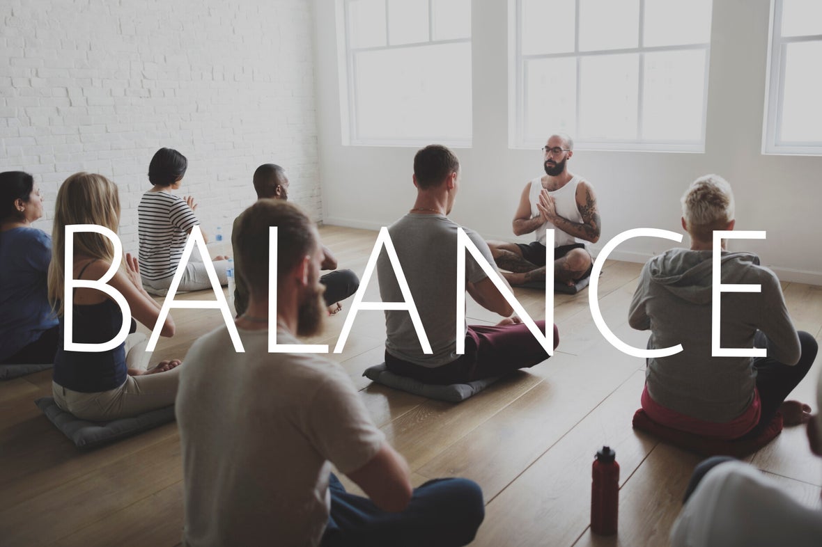 Gruppe von acht Menschen sitzt im Schneidersitz auf Yogamatten in einem hellen Raum, vorne sitzt ein Mann mit tätowierten Armen und verschränkten Händen, darüber steht das Wort 'BALANCE'