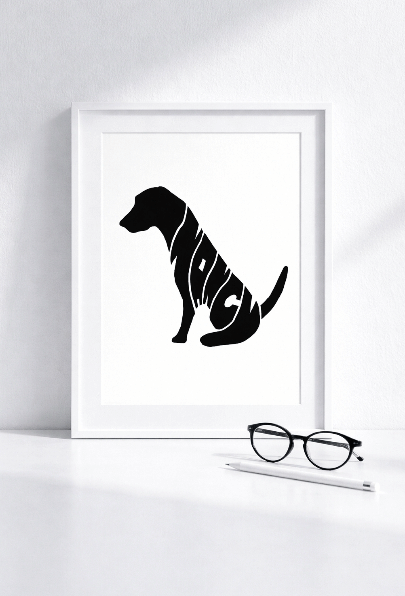 A minimalist black silhouette of a sitting dog with the name "NANCY" creatively woven into the design. The white-framed print is leaning against a white wall on a desk, accompanied by black-rimmed glasses and a pencil.