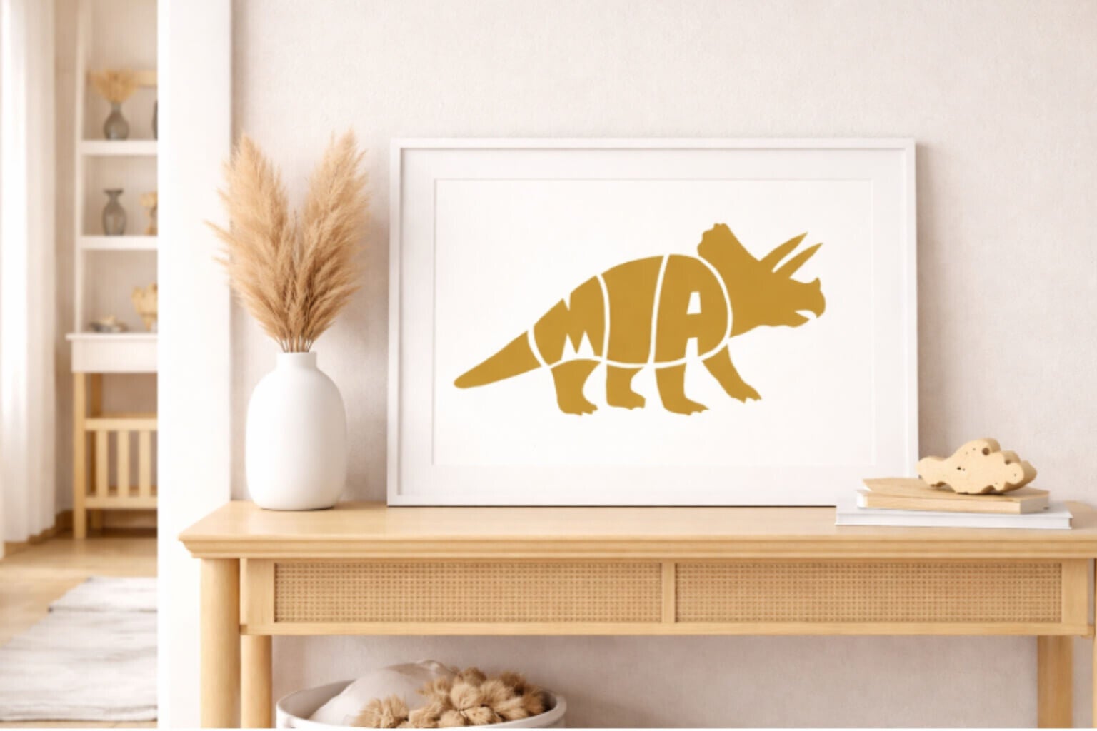 An Amber Gold typographic silhouette of a Triceratops with the name "MIA" integrated into its side. The white-framed horizontal print is displayed on a wooden sideboard in a warm, bohemian-style room next to pampas grass in a white vase.