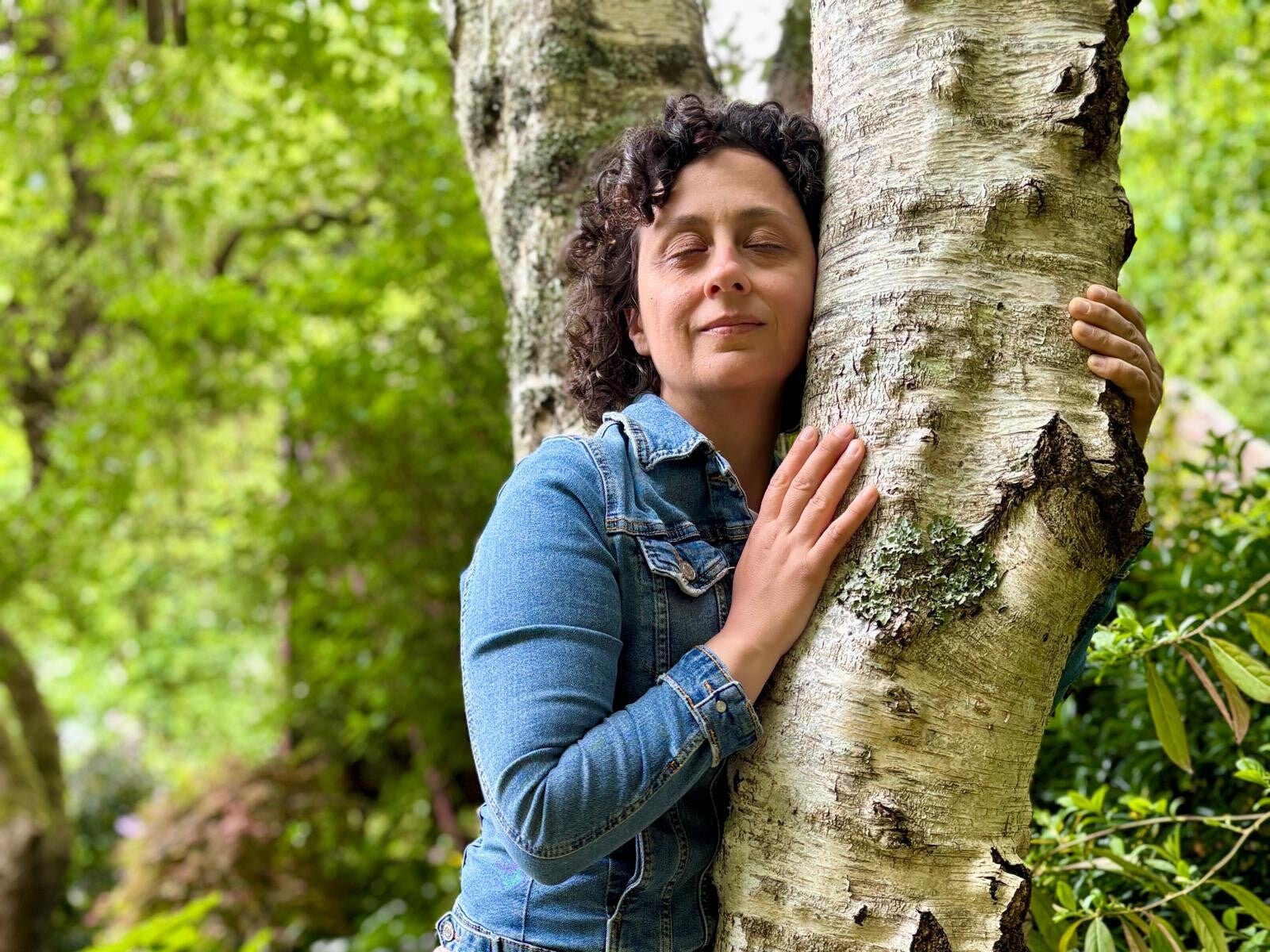Donna con gli occhi chiusi appoggiata al tronco di una betulla, immersa nella natura, in un momento di ascolto, presenza e apertura alla vita.