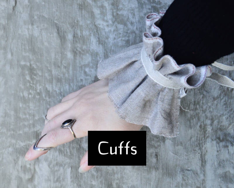 CUFFS button; afbeelding toont een bleke hand voor een rustieke grijze muur. De persoon draagt gothic ringen en een decoratief manchet uit naturel linnen