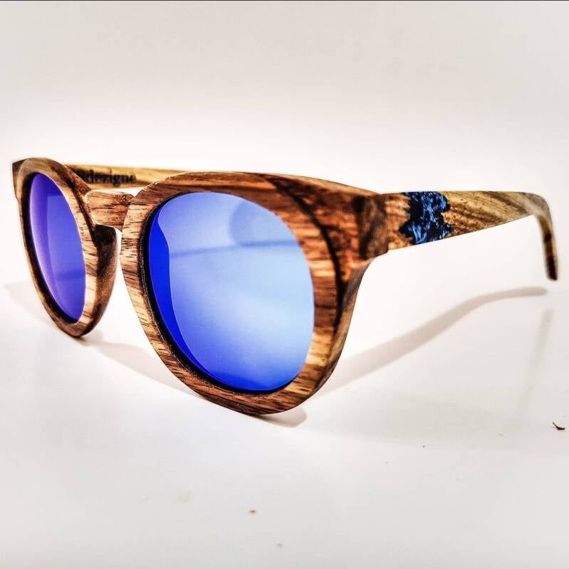 Il marchio dell'artigianato: primo piano di un paio di occhiali da sole in legno con lenti specchiate blu e l'incisione del logo sulla stanghetta.