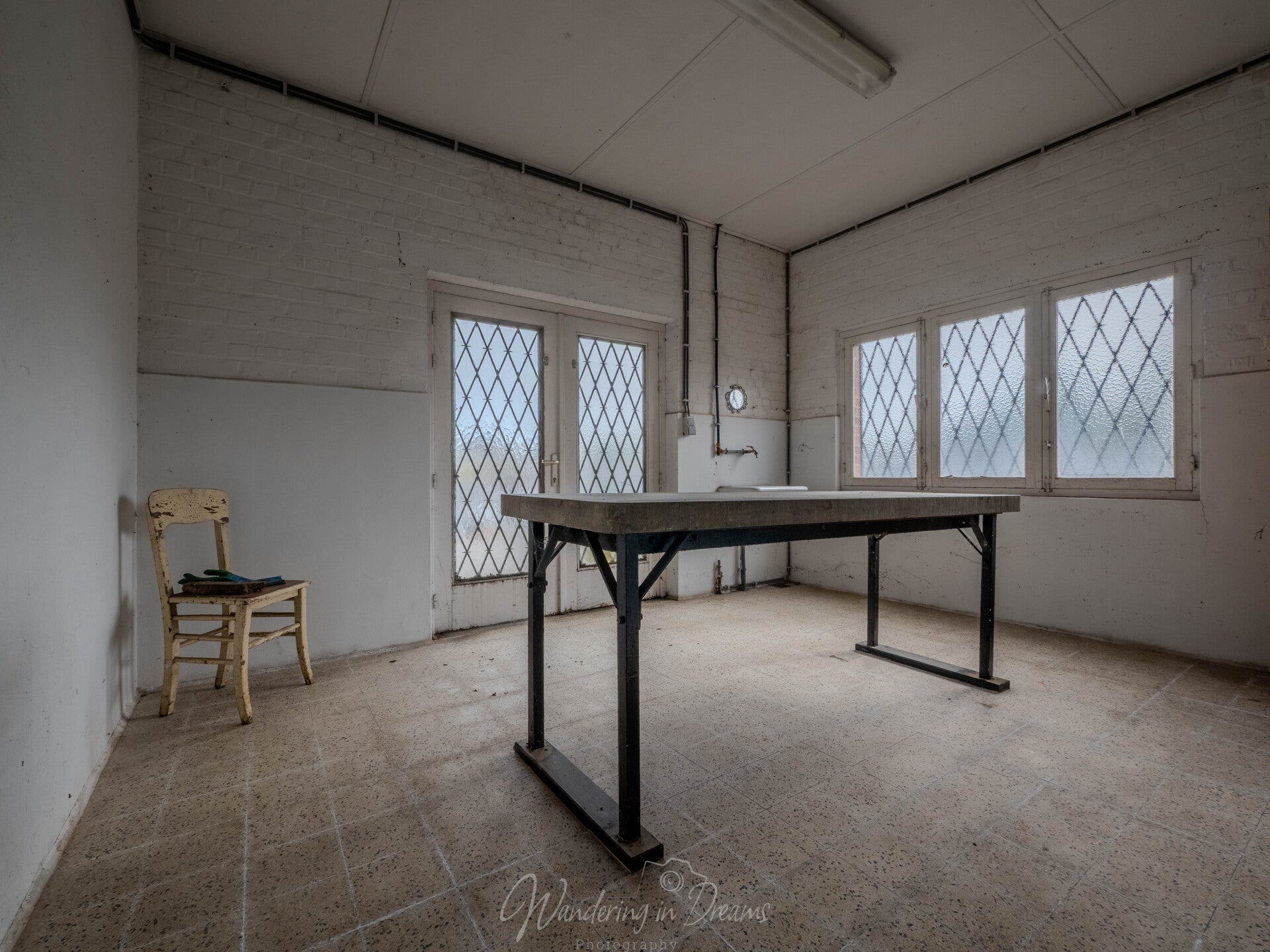Verlaten dodenhuisje op een Belgische begraafplaats, een sobere urbex locatie met lege ruimte, eenvoudige tafel en stoelen en tl-verlichting, waar de kille rust en minimalistische inrichting de functie van het mortuarium voelbaar maken