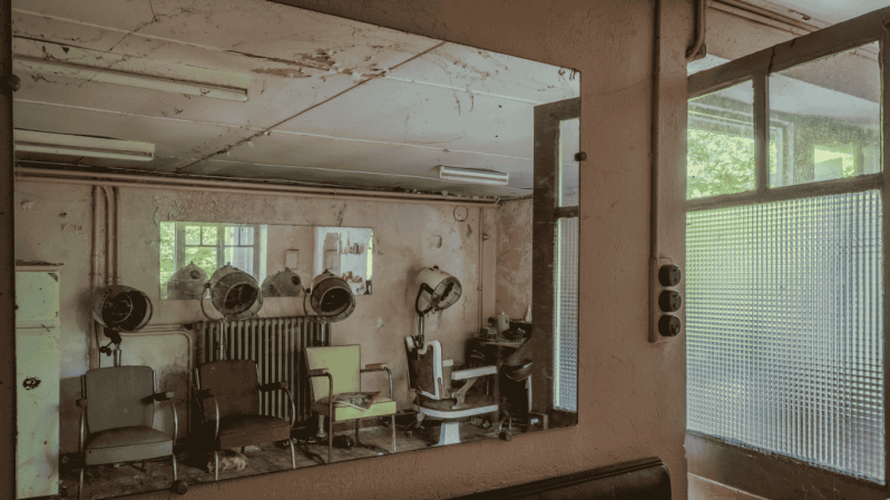 Vintage kapsalon in een verlaten Frans gebouw met retro droogkappen en gekleurde stoelen. De reflectie in de grote spiegel toont de stoffige, verstilde sfeer van dit "salon de coiffure" in Frankrijk.