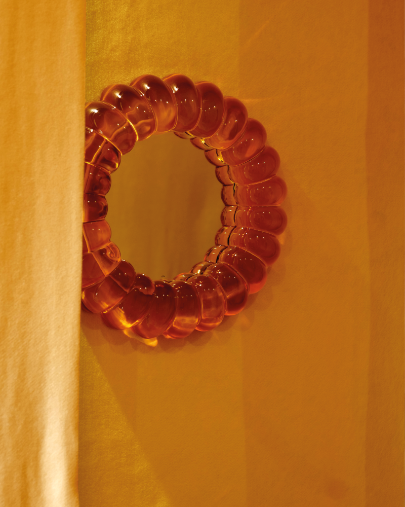 Amber orange glass mirror in a warm yellow-orange interior, showing glossy translucent material, rounded form and atmospheric colour inspiration for interiors and home decor.
