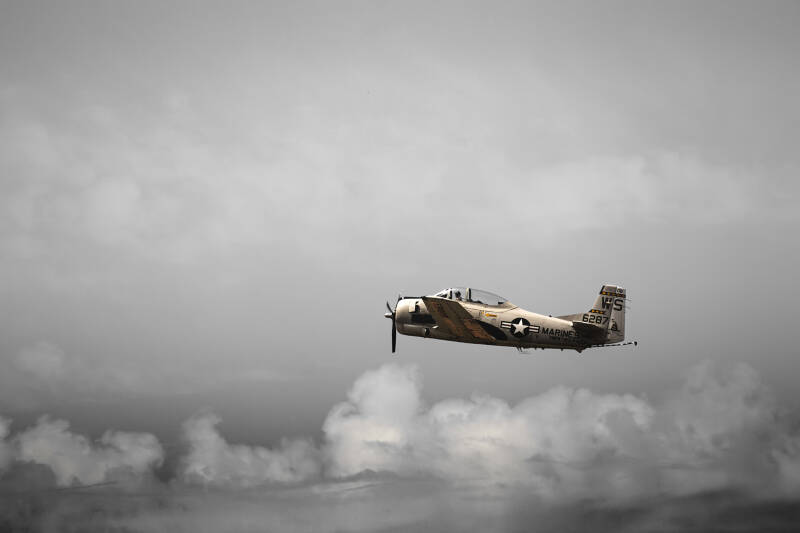 Avion North American T-28C capturé en vol au Salon du Bourget 2025, photographie en noir et blanc partiel avec accent de couleur sur l'appareil, ciel nuageux en arrière-plan.