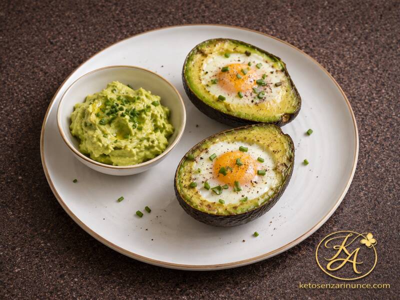 Avocado al forno con uovo cotto direttamente nella cavità, servito con cracker keto e una ciotolina di crema di avocado preparata con la polpa tolta, in una presentazione elegante perfetta per il blog.