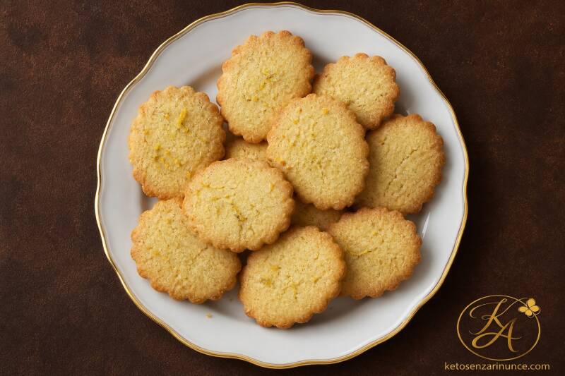 Biscotti chetogenici al limone e rum preparati con farina di mandorle, friabili e profumati, serviti su piatto bianco con bordo dorato e sfondo marrone del blog ketosenzarinunce.com.
