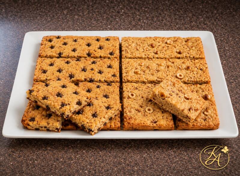 Biscotti keto in teglia tagliati a quadrotti, metà con gocce di cioccolato e metà con nocciole, disposti su un elegante vassoio bianco orizzontale su sfondo marrone speckled con logo Keto Amica.