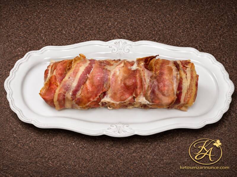 Bon roll chetogenico al forno avvolto nello speck, sistemato su un elegante vassoio bianco su sfondo marrone speckled con logo KA e scritta ketosenzarinunce.com in basso a destra. Immagine blog pulita e raffinata, adatta a ricetta keto salata.