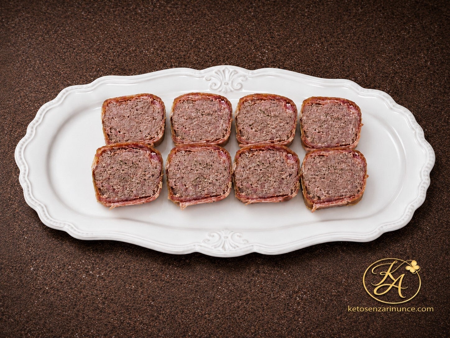 Fette di bon roll chetogenico avvolto nello speck, disposte su un elegante vassoio bianco orizzontale su sfondo marrone speckled con logo KA e scritta ketosenzarinunce.com in basso a destra. Immagine pulita e raffinata, adatta al blog per presentare il bo