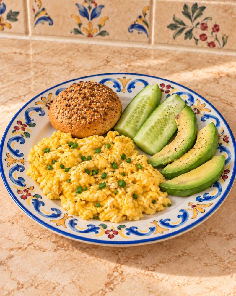 Colazione chetogenica salata con focaccina keto alla ricotta e mortadella, uova strapazzate con erba cipollina, avocado e cetriolo fresco, serviti su un piatto mediterraneo decorato.