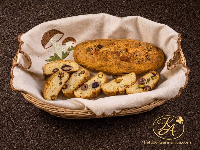 Filoncini keto alla crescenza con olive taggiasche e pistacchi, presentati in un elegante cesto di vimini con alcune fette tagliate che mostrano la mollica compatta e morbida. Pane chetogenico rustico, profumato e con una crosta dorata