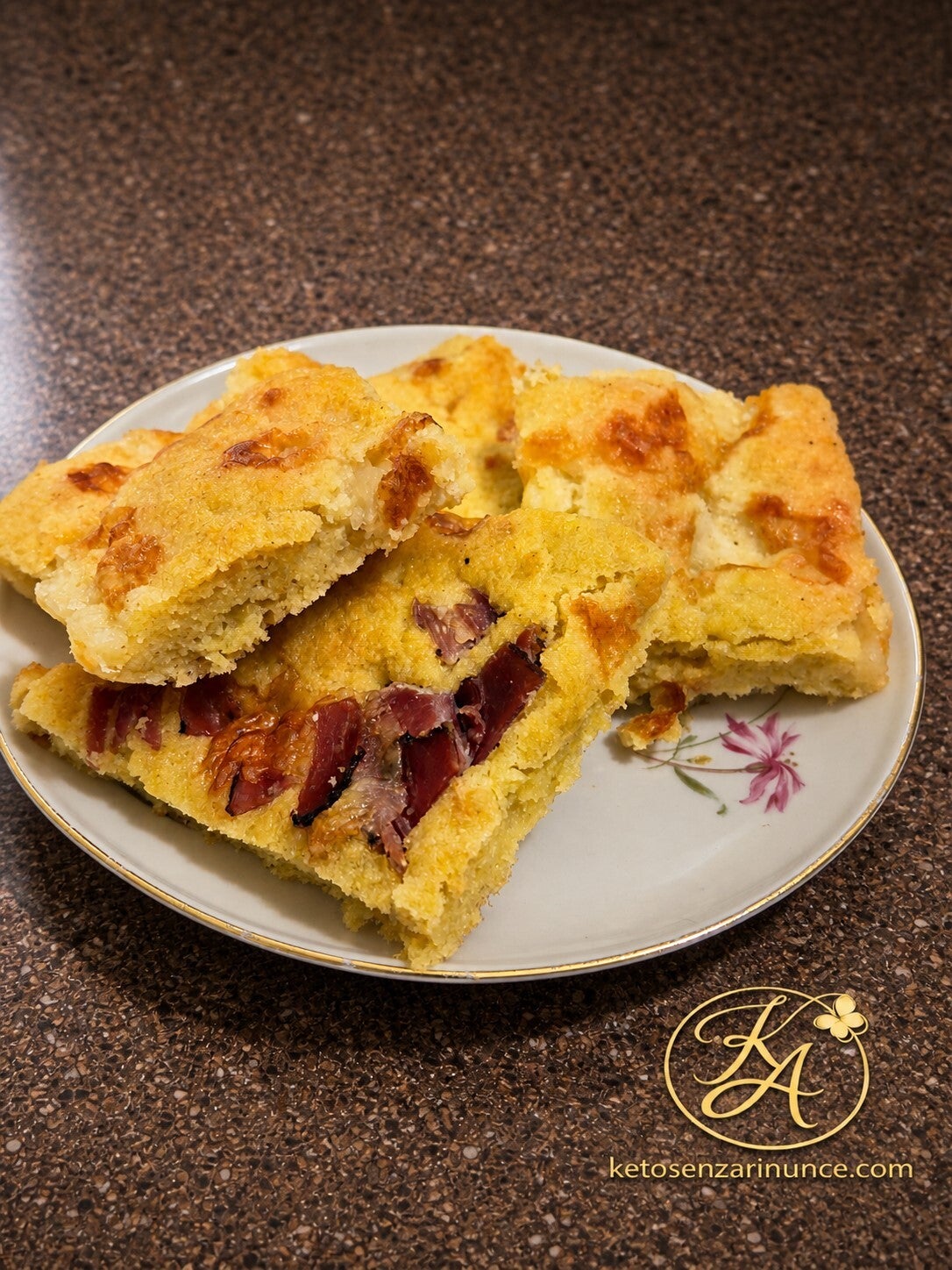 Focaccia salata keto con prosciutto e formaggio, tagliata a quadrotti morbidi e dorati, servita su piatto chiaro con bordo dorato sullo sfondo blog di ketosenzarinunce.com.