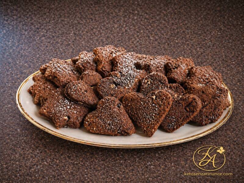 Frollini keto al doppio cioccolato con noci e arancia, disposti su un piatto bianco con bordo dorato, fotografati su sfondo marrone speckled con logo Keto senza rinunce.