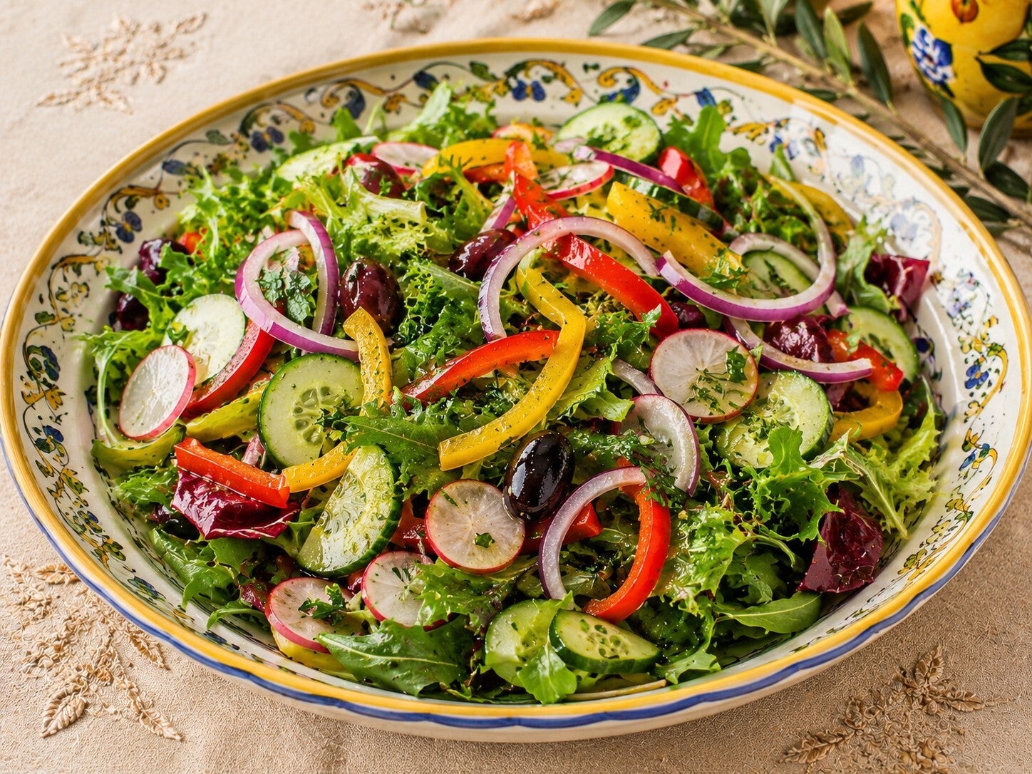 Insalata mediterranea colorata con foglie verdi, cetrioli, ravanelli, peperoni rossi e gialli, cipolla di Tropea e olive, servita in una ciotola decorata su tovaglia beige ricamata.