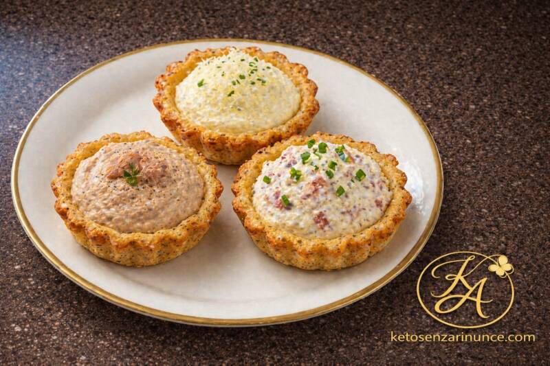 Tre mini crostatine salate chetogeniche servite su piatto bianco con bordo oro. Le crostatine sono farcite con crema al formaggio, patè di tonno e crema di ricotta e speck. Idea perfetta per antipasto, buffet o colazione salata keto