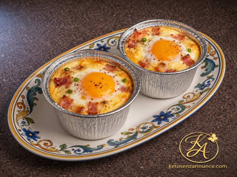 Due mini tortini keto di uova e pancetta cotti al forno in stampini monoporzione di alluminio, serviti su un vassoio ovale bianco con bordi decorati in stile mediterraneo. Una colazione salata chetogenica semplice, saziante e scenografica, perfetta per in