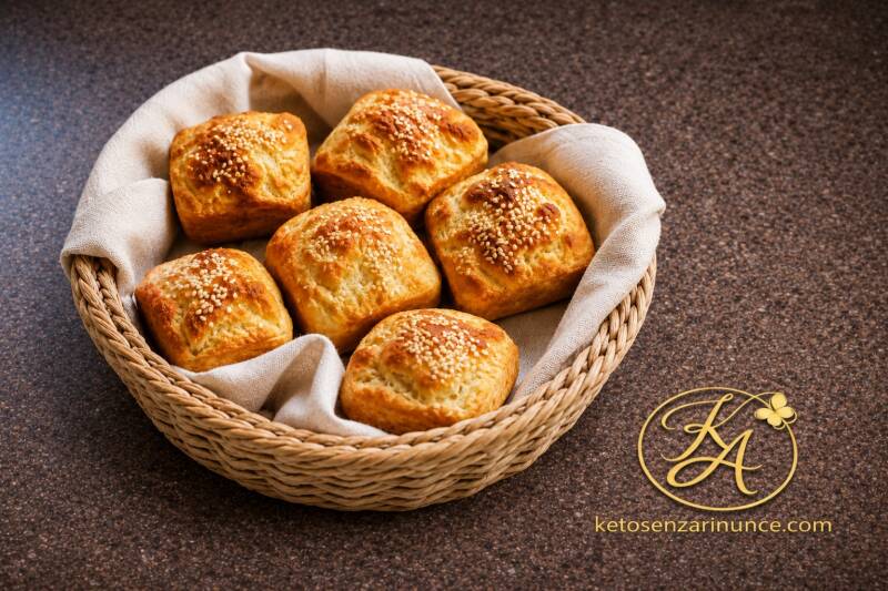 Panini keto al sesamo dorati, disposti in un cestino di vimini rivestito con telo chiaro, su sfondo marrone speckled con logo KA dorato e riferimento a ketosenzarinunce.com. Immagine calda, pulita ed elegante per il blog Keto senza rinunce, ideale per pre
