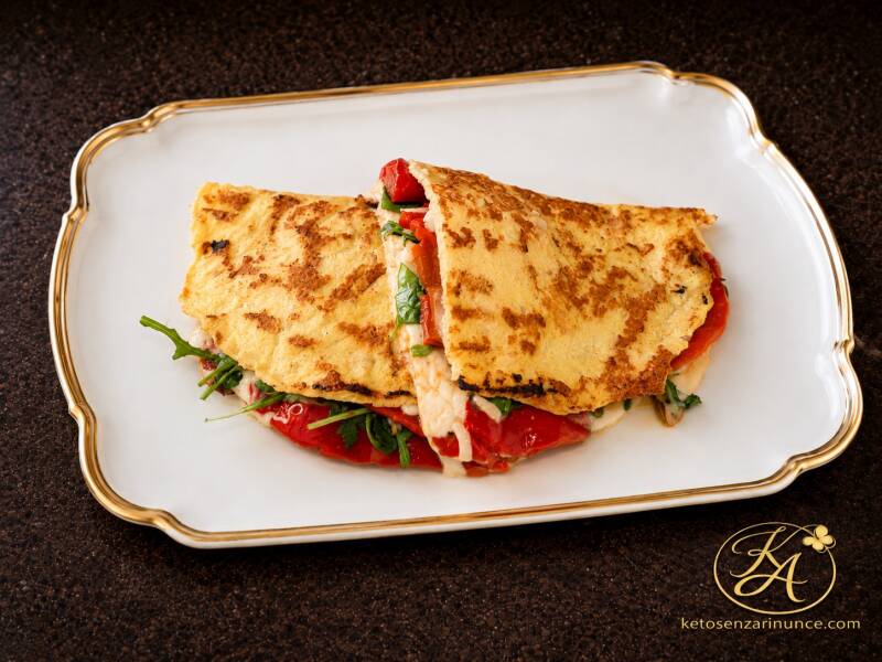 Piadina keto di petto di pollo farcita con peperoni al forno, scamorza e rucola, servita su vassoio bianco con bordo dorato su sfondo marrone del blog ketosenzarinunce.com. Una ricetta proteica, senza farina e senza frittura, perfetta per un pranzo o una 