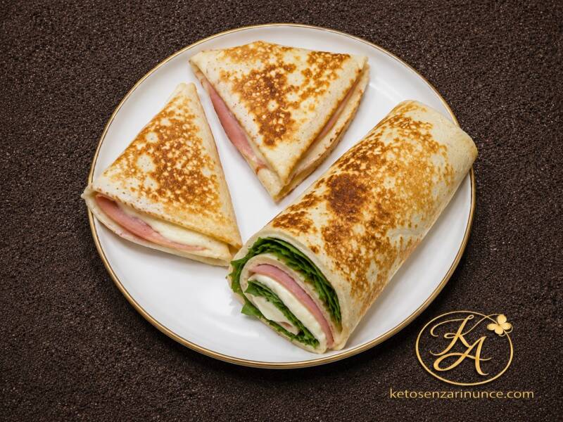 Tre piadine keto senza farina di mandorle con albumi, servite su piatto bianco con bordo oro: due piegate a triangolo e una arrotolata, farcite con prosciutto, formaggio e insalata.