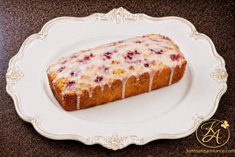 Plumcake keto alla panna e lamponi soffice e profumatissimo, servito intero su piatto bianco elegante con velo di glassa chiara, su sfondo marrone speckled con logo KA e riferimento a ketosenzarinunce.com. Immagine dolce raffinata per il blog Keto senza r