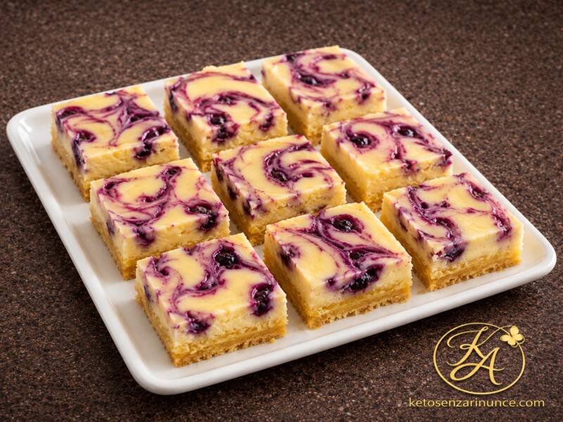Pasticcini keto al limone e mirtilli disposti su un vassoio bianco, con superficie chiara marmorizzata ai frutti di bosco e sfondo marrone speckled con logo ketosenzarinunce.com.