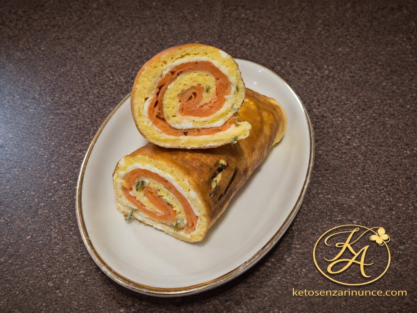 Roll di uova al forno farcito con salmone e formaggio cremoso, presentato tagliato a metà su piatto bianco con bordo dorato, in uno stile semplice ed elegante perfetto per il blog.